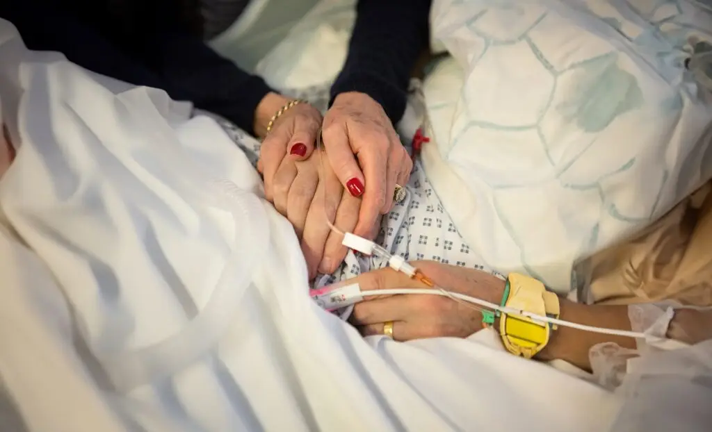 Close-up of two pairs of hands on a hospital bed. One pair of hands has red fingernails and is clasped. The other visible hand has a drip in it
