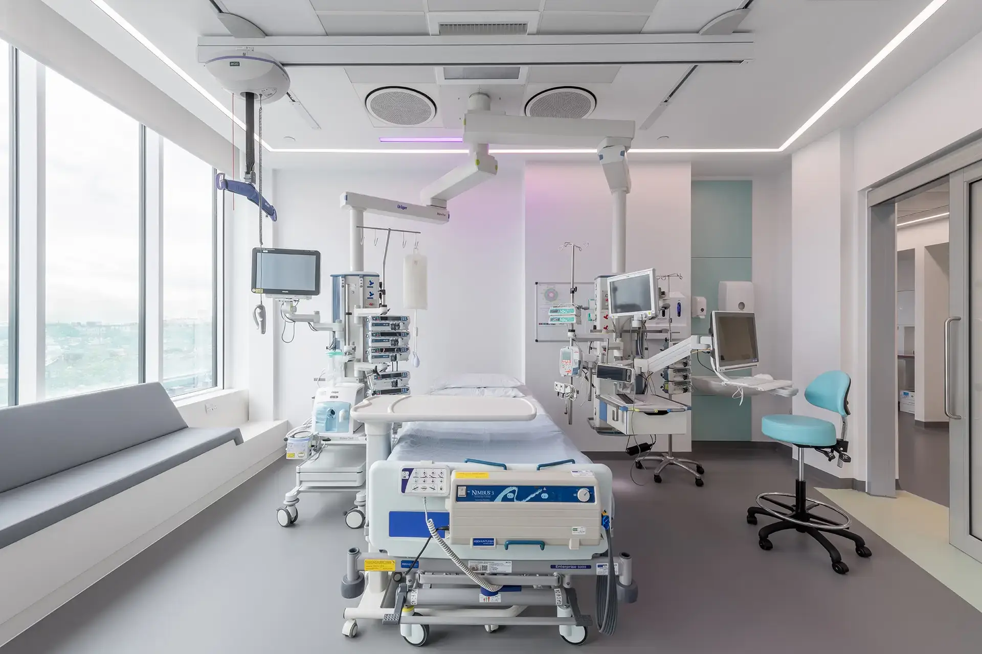 Hospital room with large windows, a window seat and a hospital bed with lots of equipment around the bed. It is a modern-looking space