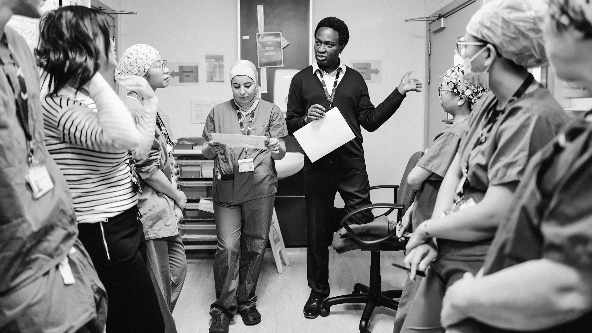 Photo by James Kent of a team meeting at West Middlesex University Hospital. Staff members are standing in a group listening to a consultant briefing them