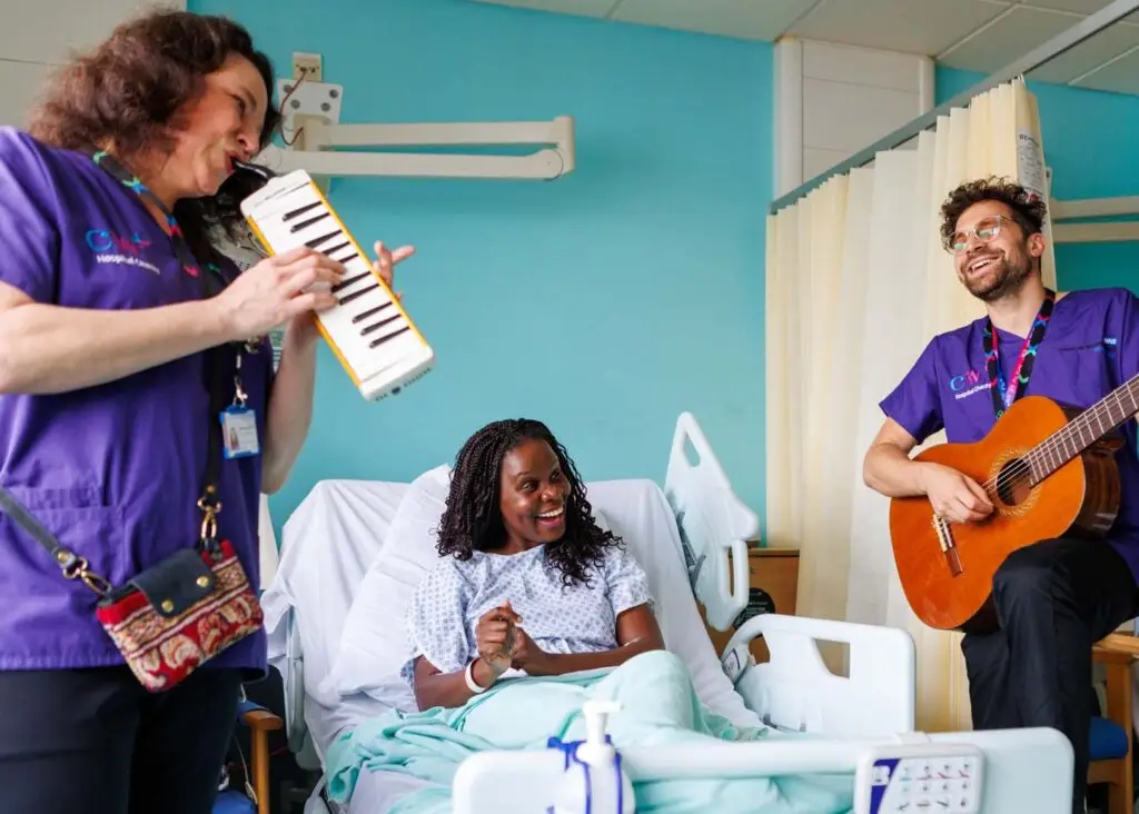 Smiling Black woman in a hospital bed. There are two musicians performing at her bedside: on one side, a woman playing a melodica and on the other, a man playing a guitar