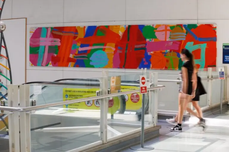 Two people about to get on a descending escalator, with a large, colourful artwork on the wall behind them by Albert Irvin