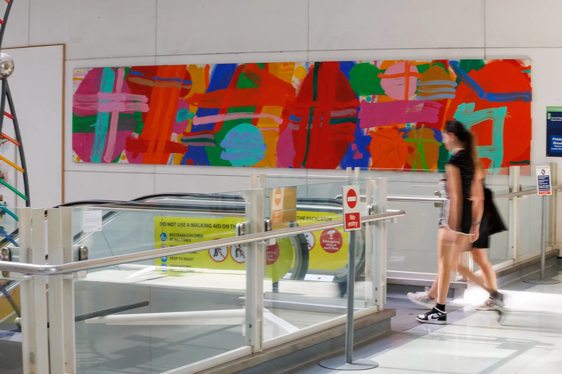 Two people about to get on a descending escalator, with a large, colourful artwork on the wall behind them by Albert Irvin