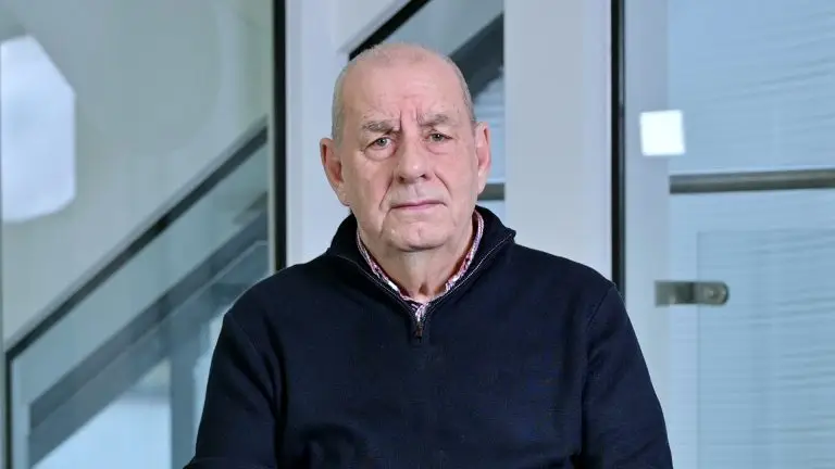 A serious-looking man with a bald head wearing a black jumper. He is being interviewed about a cancer scare
