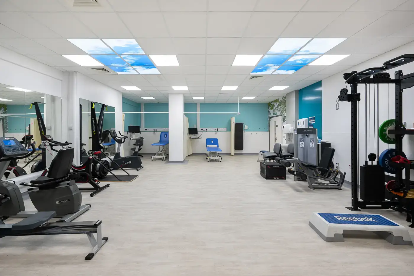 Large gym with various pieces of equipment and LED ceiling tiles depicting blue sky and clouds