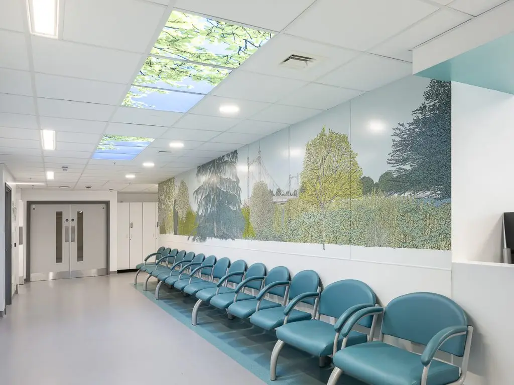 Hospital corridor with blue seats, wall art depicting a nature scene with trees, and LED ceiling tiles with leaves and blue sky