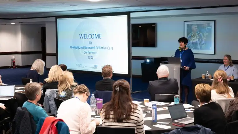 Speaker presenting to an audience with a slide that reads 'WELCOME to the National Neonatal Palliative Care Conference 2025'