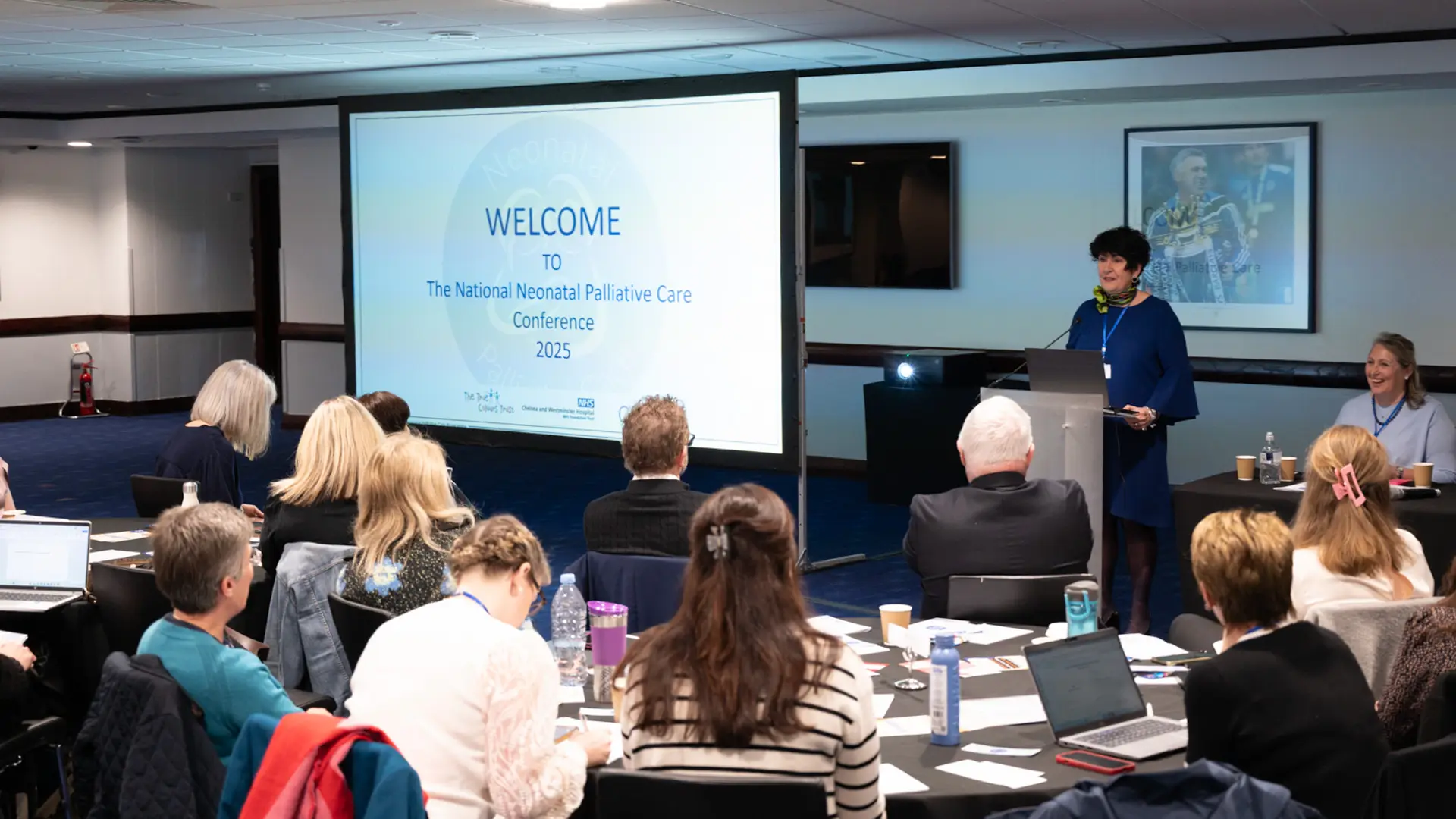 Speaker presenting to an audience with a slide that reads 'WELCOME to the National Neonatal Palliative Care Conference 2025'