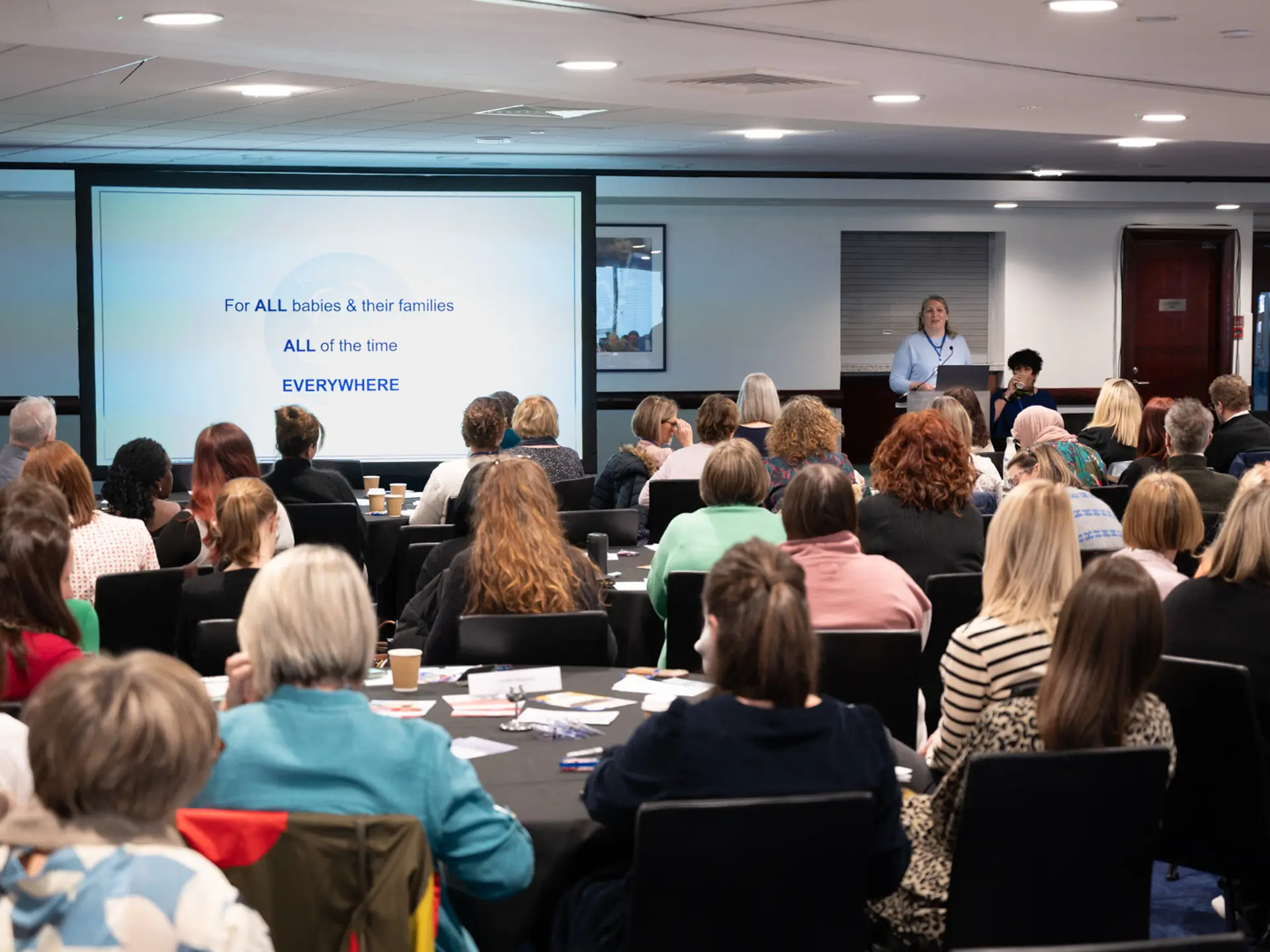 Speaker (Alex Mancini) in a conference room speaking to an audience, with a slide that reads 'For ALL babies and their families, ALL of the time, EVERYWHERE'