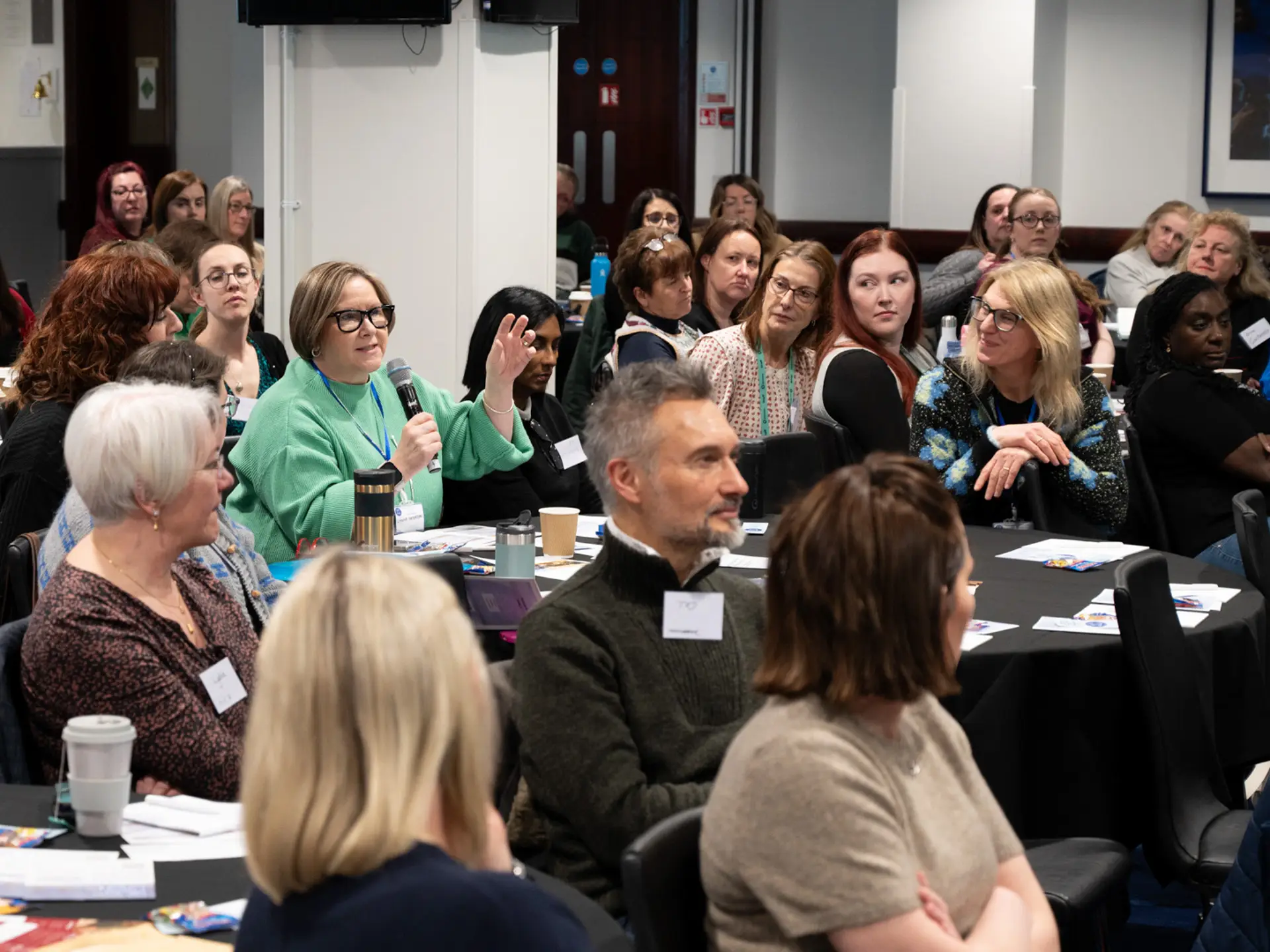 Audience in a room, with one lady wearing a green top holding a microphone to ask a question or share her expertise. This is a Neonatal Palliative Care Conference held at Chelsea Football Club