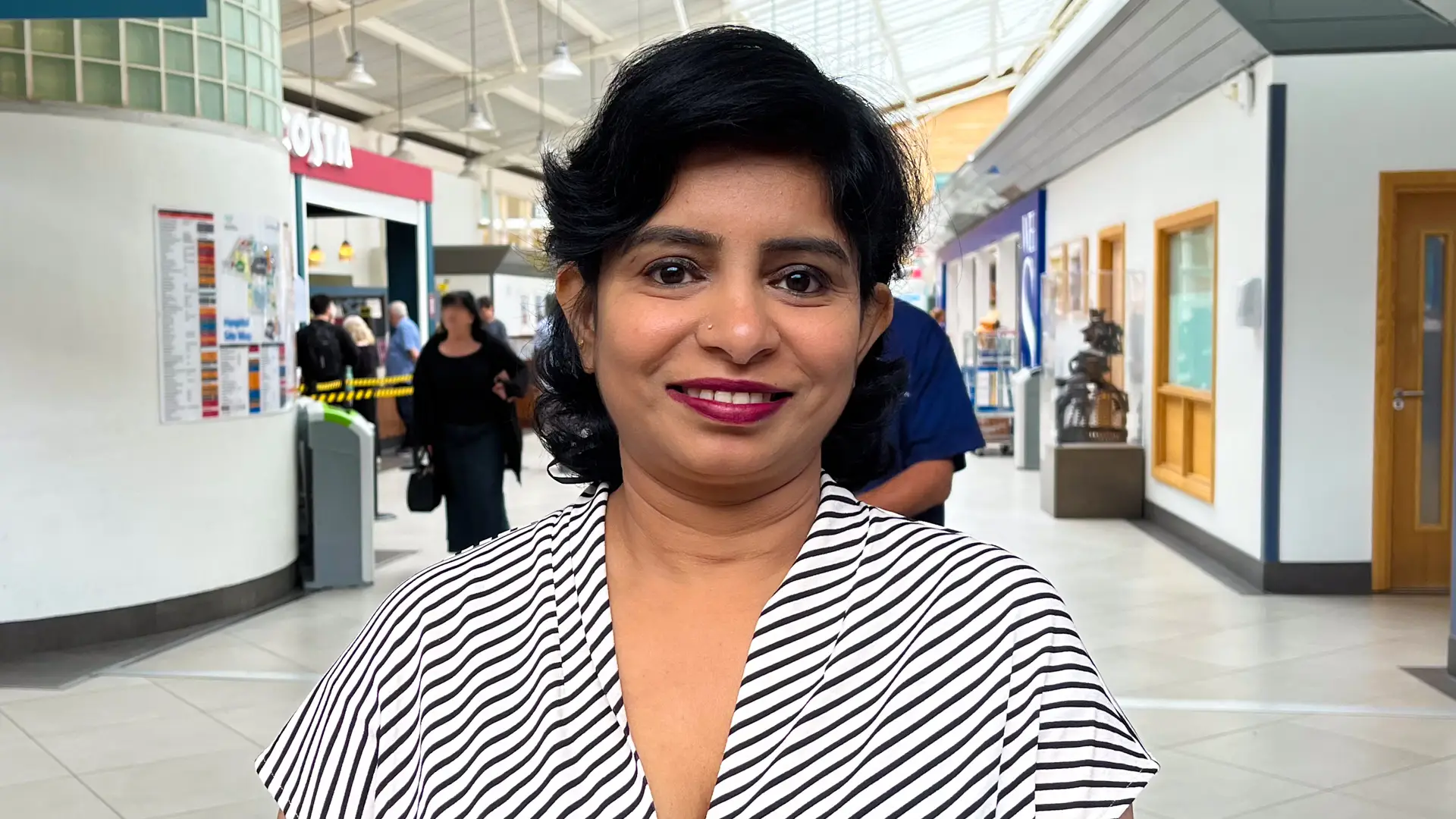 A woman with short dark hair standing in the busy atrium at West Middlesex University Hospital wearing a black and white striped top, smiling at the camera. She is helping to raise awareness of the Ambulatory Diagnostic Centre
