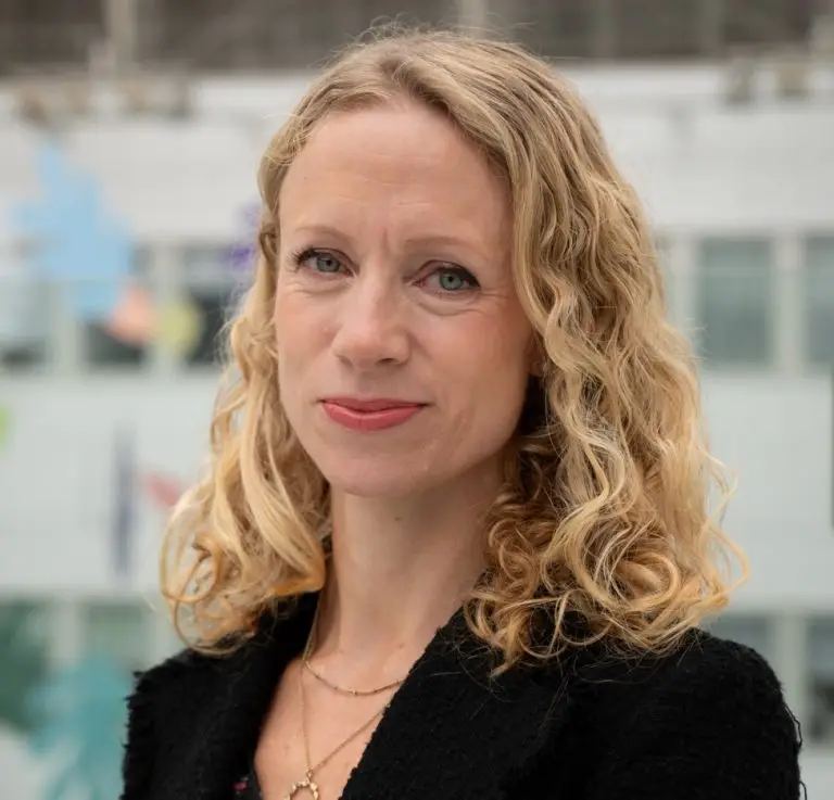 A woman with blond, slightly wavy hair looking to camera with a slight smile