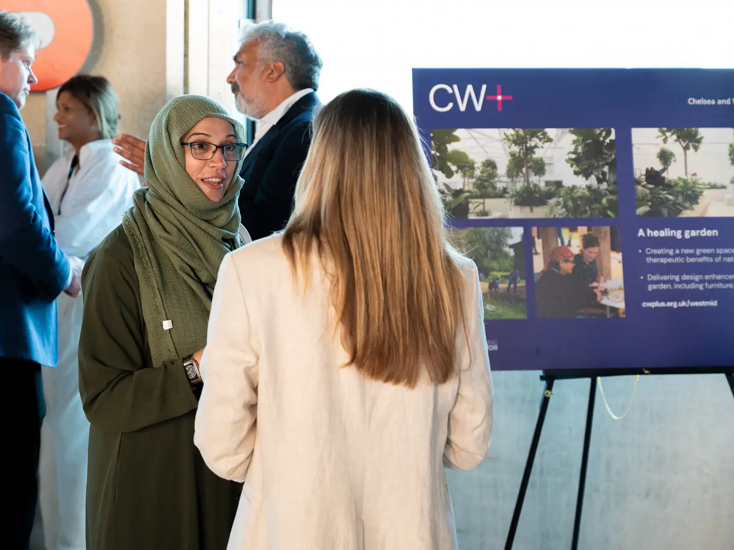 Two women chatting at an event. One is seen from behind and has long fair hair; the other is wearing glasses and a head scarf. In the background there is a CW+ information board on an easel