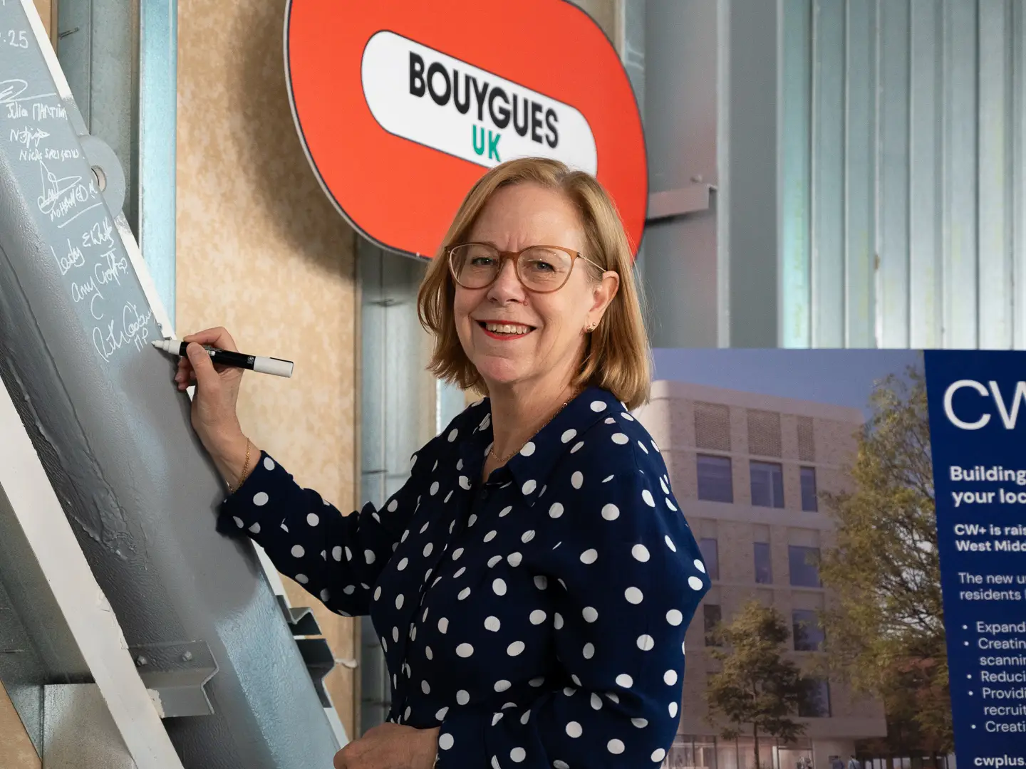 Woman (Ruth Cadbury MP in polka dot dress smiling at the camera. She is holding a pen to sign a steel beam in the new ADC building being constructed at West Middlesex University Hospital