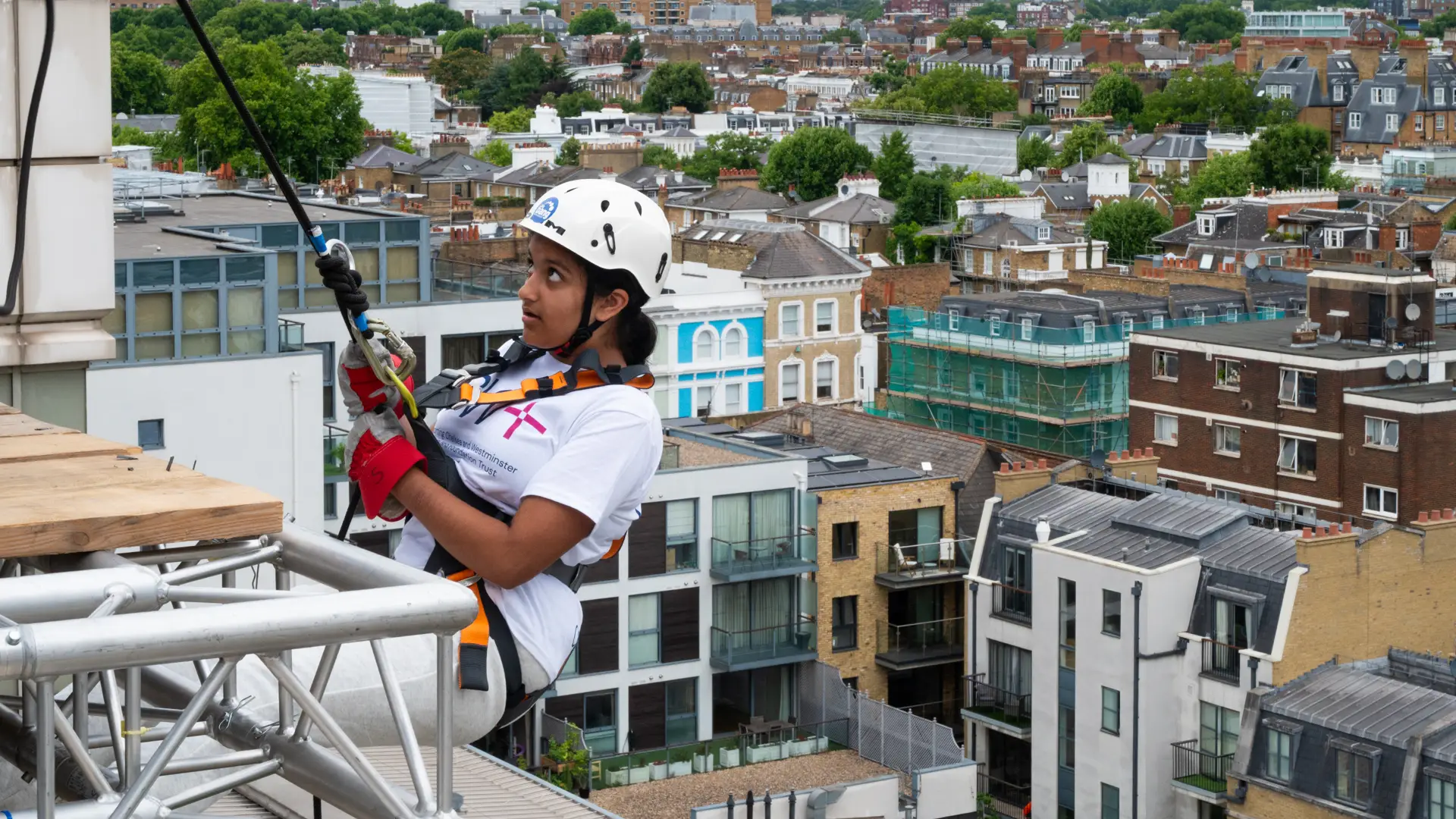 A young girl in a harness and helmet leans off the top of a building, about to start an abseil descent