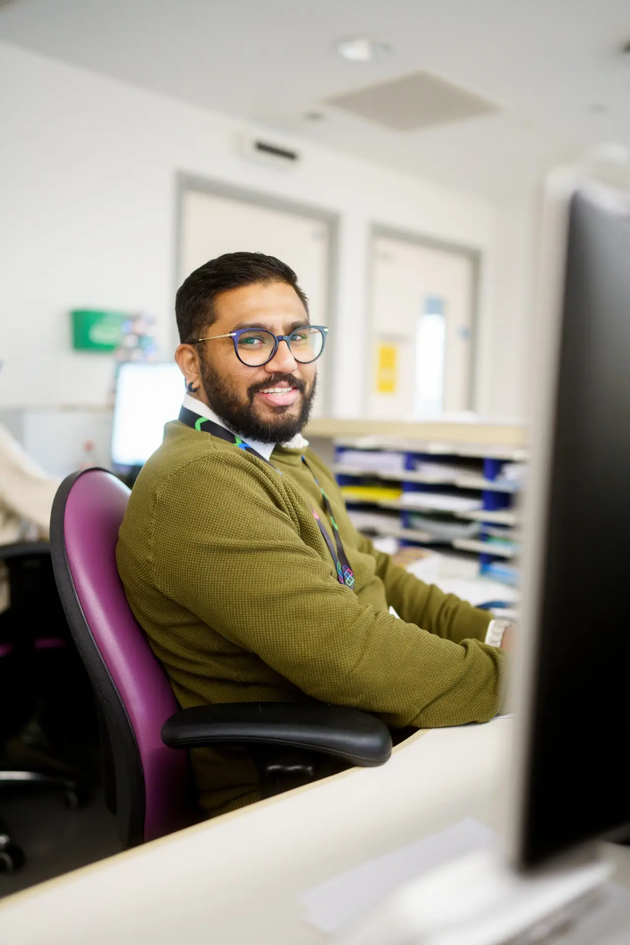 Man with a beard, glasses and an olive green jumper sitting at a work desk, smiling at the camera