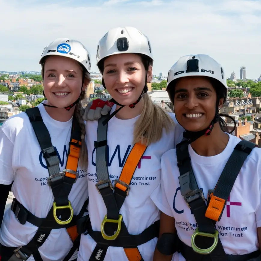 Three young women wearing hard hats, harnesses and CW+ T-shirts, smiling at the camera. They are preparing to abseil down Chelsea and Westminster Hospital