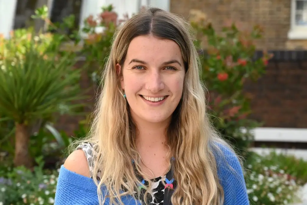 Drawn in Residence programme: A woman (Emily Thomas) with long blond hair, smiling at the camera. She is standing in front of some greenery. She is the Arts Manager at West Middlesex University Hospital and runs the Drawn in Residence programme