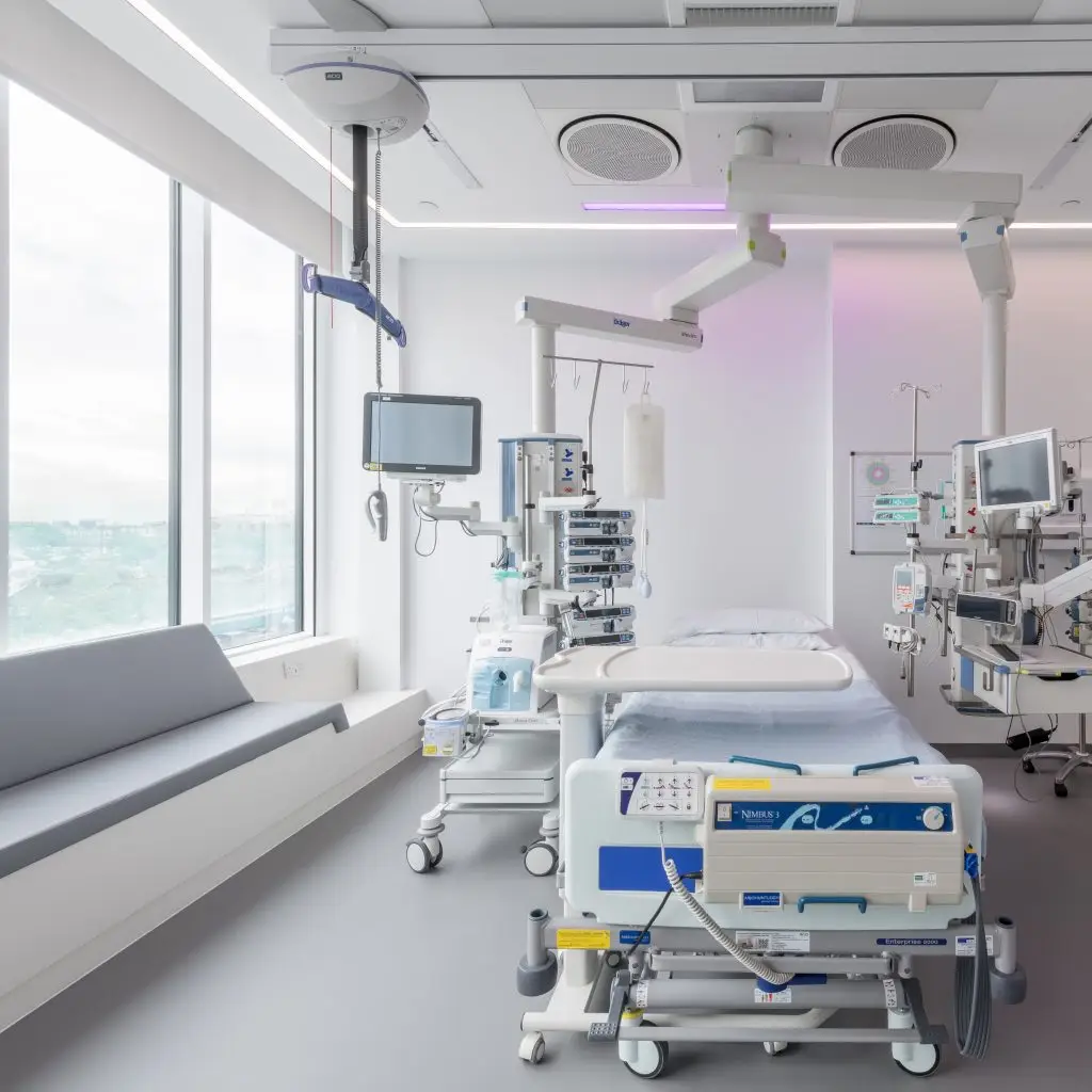Light, bright hospital room with large windows and window seating. The bed has equipment and screens around it