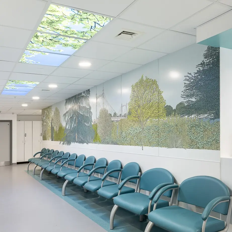 Hospital corridor with blue seating and a long mural with trees and greenery. In the ceiling there are lightboxes with sky and leaves