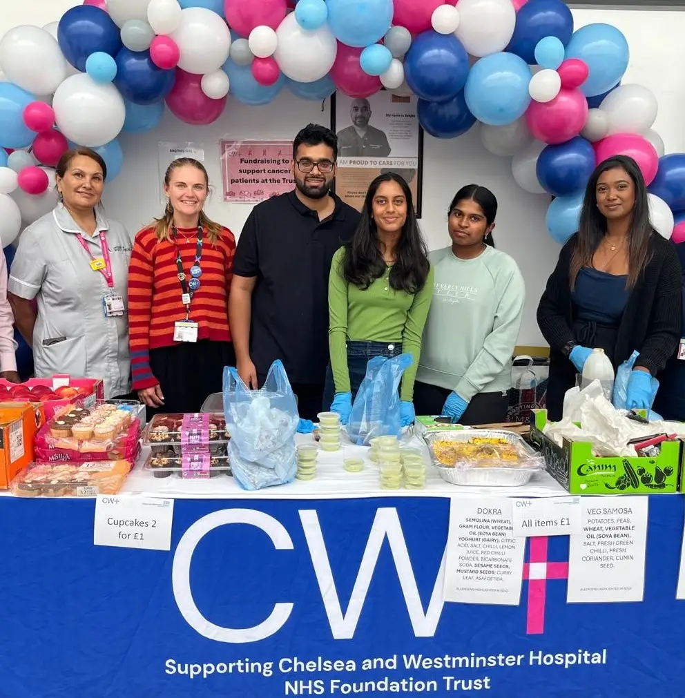 A group of people standing at a table with a CW+ branded tablecloth. There are baked goods on the table and a balloon arch over their heads