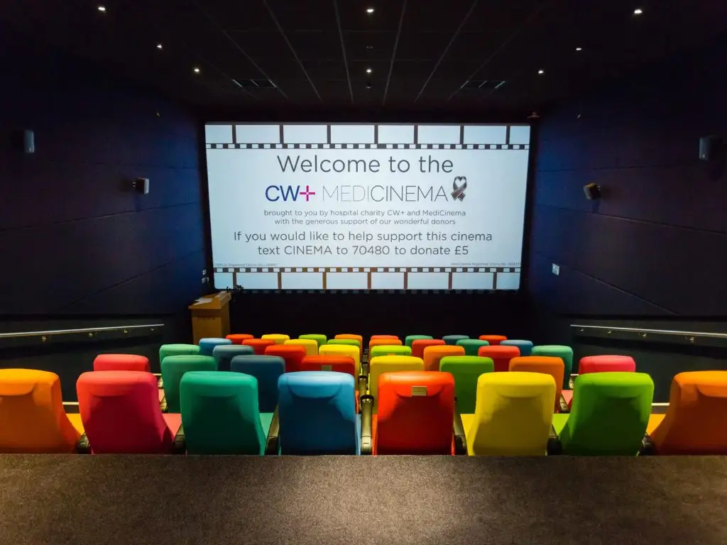 CW+ MediCinema, taken from the back of the cinema. There are raked seats in red, green, blue and yellow, The screen is showing a 'Welcome to the CW+ MediCinema' message with a text-to-donate number