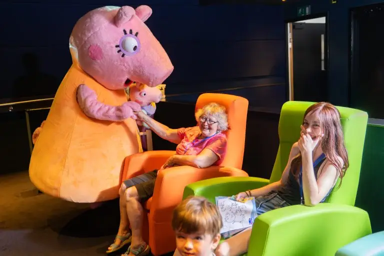 Mummy Pig (from Peppa Pig) meeting two people - an older woman sitting in an orange armchair, and a younger person sitting in a green armchair. In the foreground is a toddler. the location is CW+ MediCinema at Chelsea and Westminster Hospital