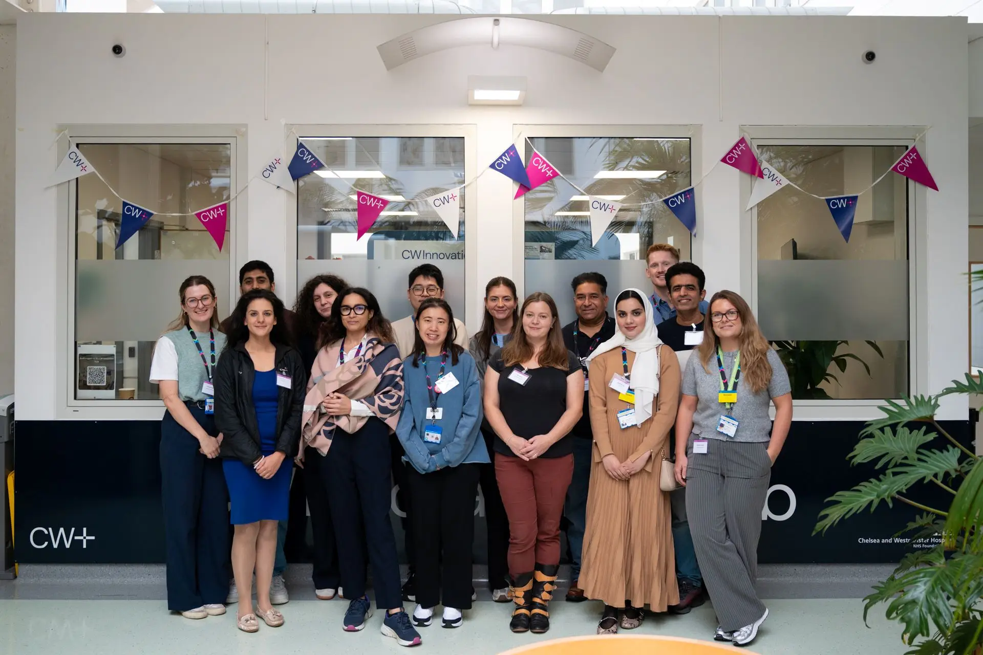A diverse group of people posing for a group photo. Behind them is some bunting that says 'CW+. They are this year's cohort of staff from Chelsea and Westminster Hospital NHS Foundation Trust taking part in the CW Innovation Fellowship