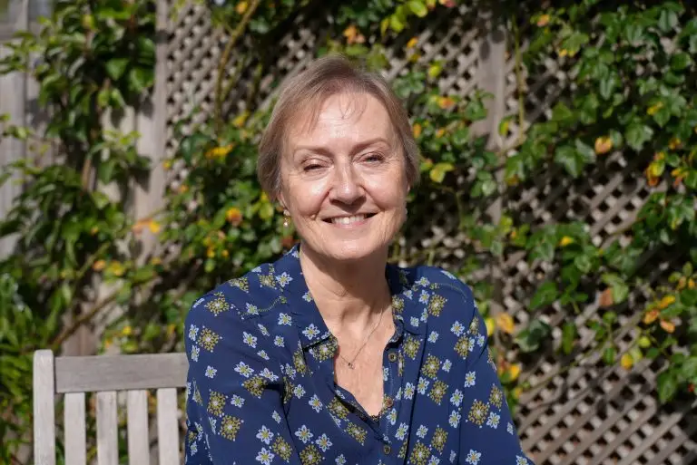 Linda's story: White woman wearing a blue shirt with a white motif, smiling at the camera. She is outside, and behind her is a trellis with greenery. She is Linda Alexander, the focus of the Christmas campaign 2025 and the final story in our Thirty Stories series