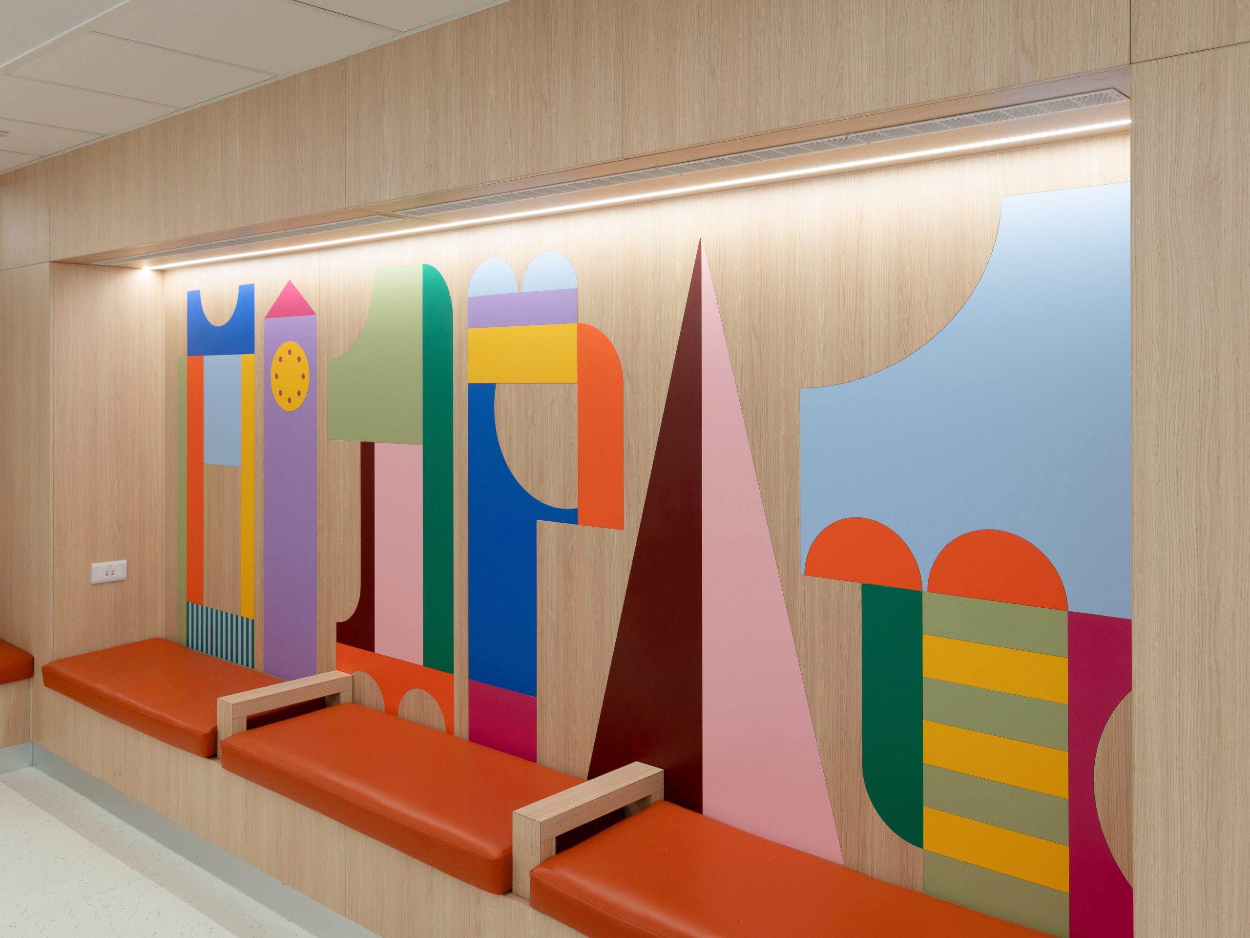 A 'nook' with padded bench seating, built-in lighting and a wall collage made out of simple blocks in bright colours. The shapes are simplified abstract depictions of London landmarks. This is in the Children's Emergency Department at Chelsea and Westminster Hospital. The art was designed by Irish artist Gráinne Nagle