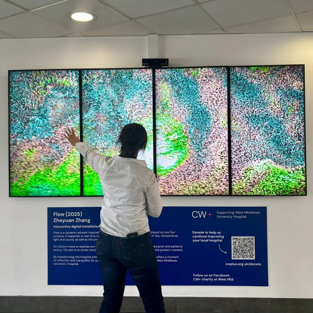 Four large screens display a colourful digital artwork with swirling shapes in blue, green and pink. A woman stands in front of it with her arm outstretched