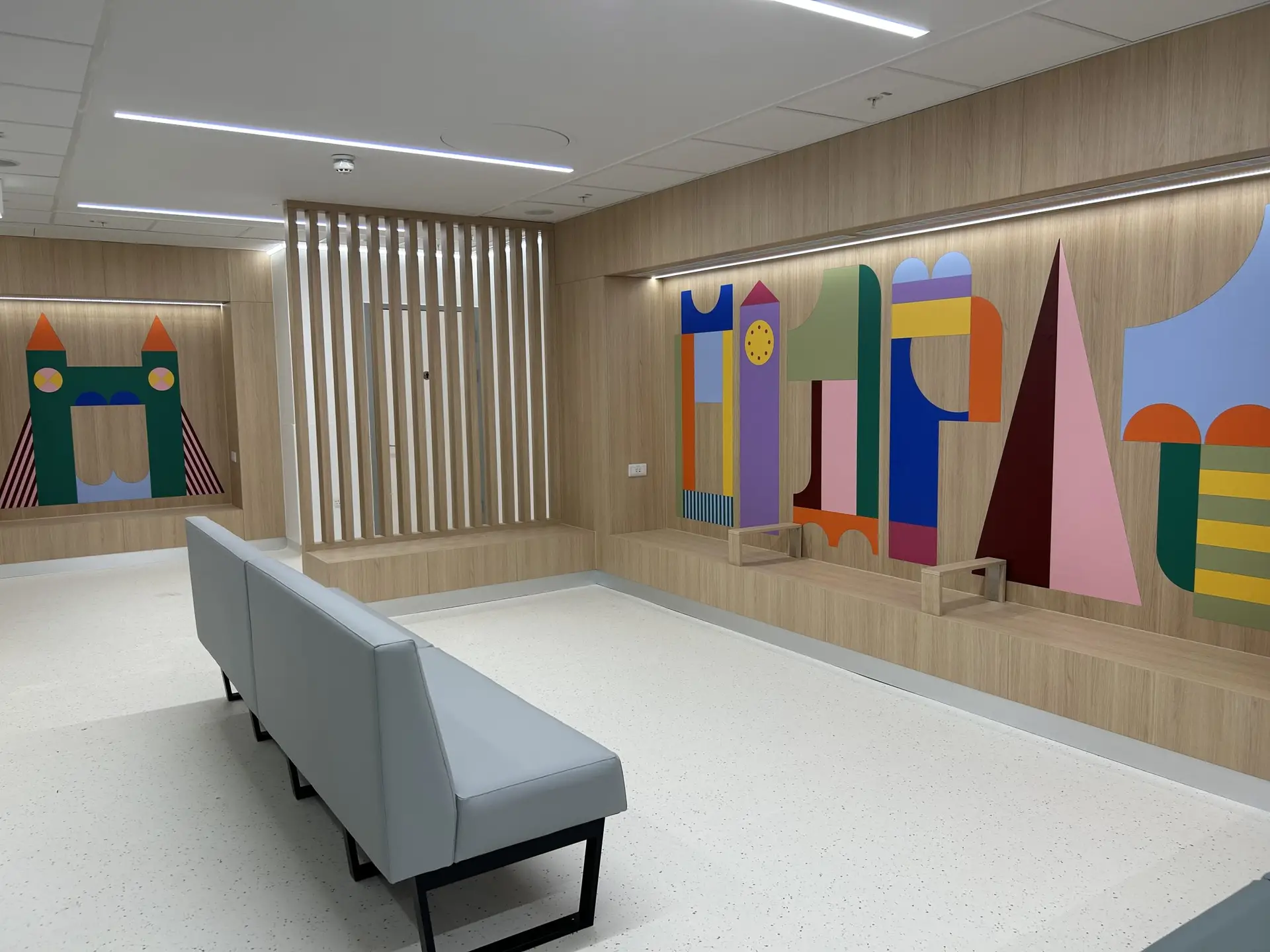 A hospital waiting area with padded bench seating facing a seating nook with a wall collage made out of simple blocks in bright colours. The shapes are simplified abstract depictions of London landmarks. This is in the Children's Emergency Department at Chelsea and Westminster Hospital. The art was designed by Irish artist Gráinne Nagle