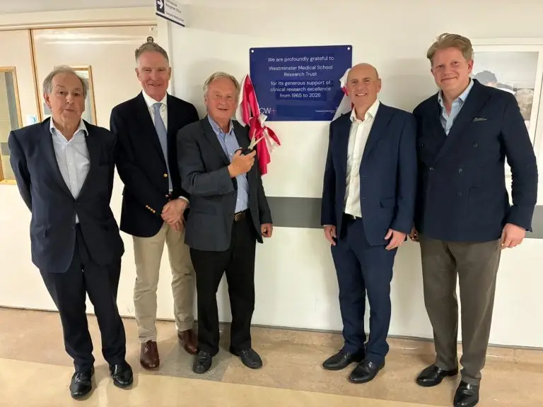 Five people stand in a corridor at Chelsea and Westminster Hospital beside a newly unveiled blue plaque on a wall. One person is holding a red ribbon used in the unveiling. The plaque reads: 'We are profoundly grateful to Westminster Medical School Research Trust for its generous support of clinical research excellence from 1965 to 2026.'