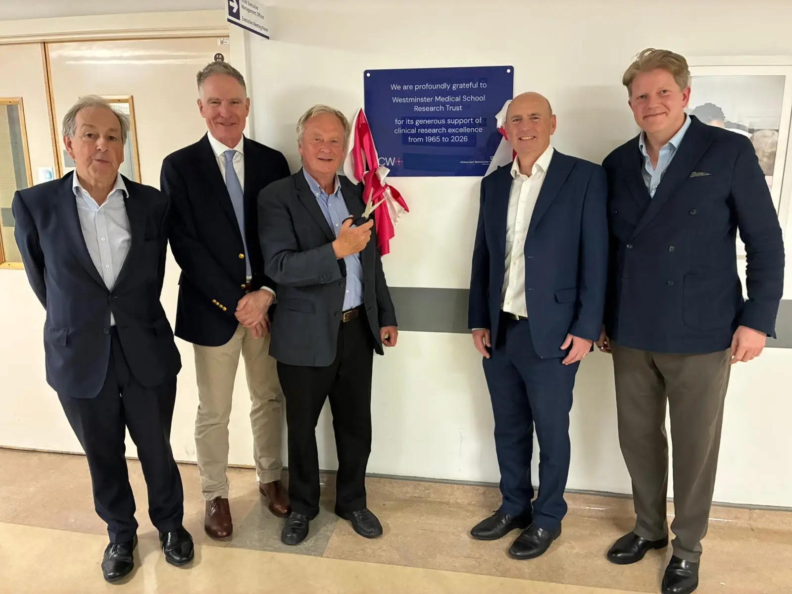 Five people stand in a corridor at Chelsea and Westminster Hospital beside a newly unveiled blue plaque on a wall. One person is holding a red ribbon used in the unveiling. The plaque reads: 'We are profoundly grateful to Westminster Medical School Research Trust for its generous support of clinical research excellence from 1965 to 2026.'
