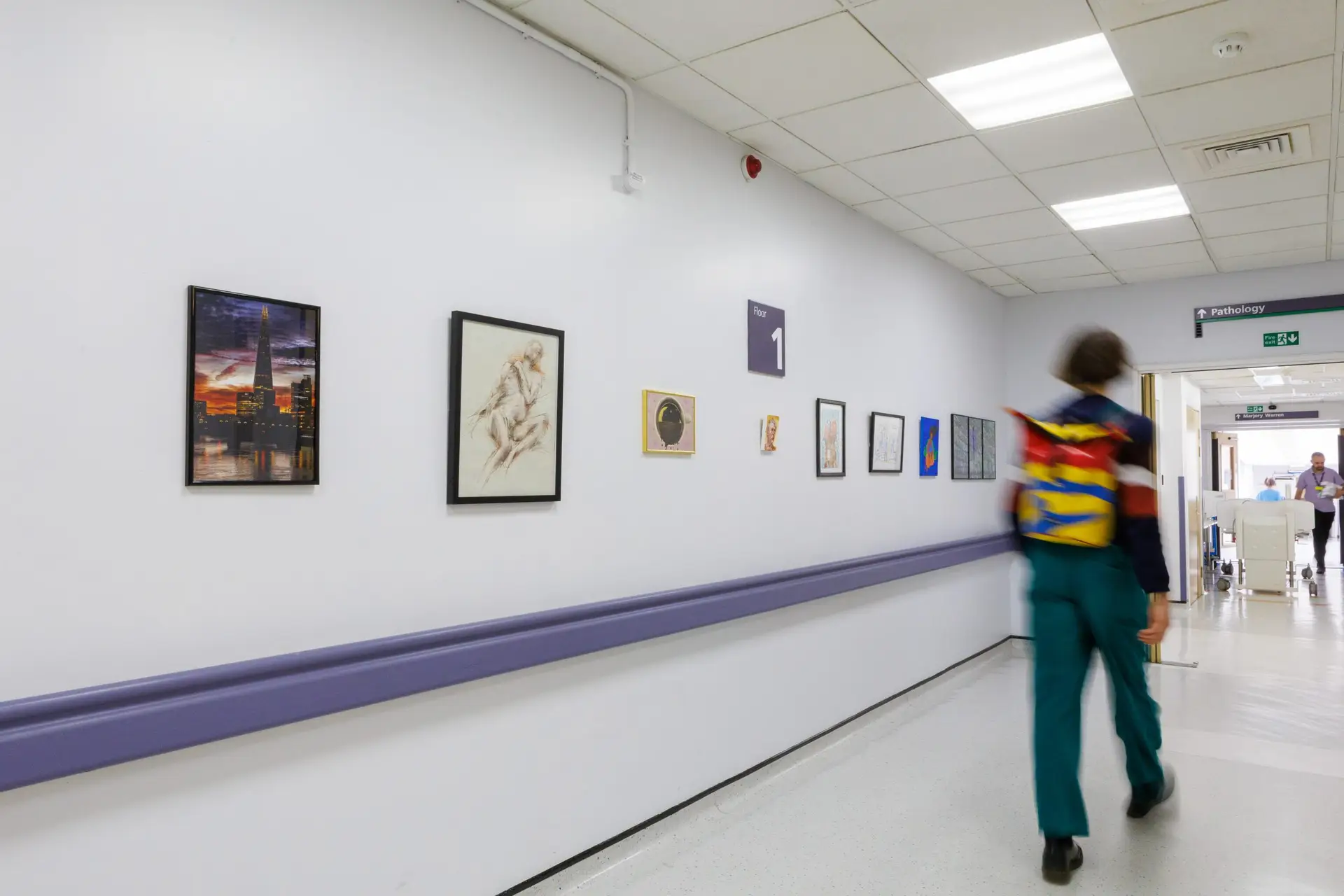 A gallery of art on a white wall. A man with a colourful backpack is walking past