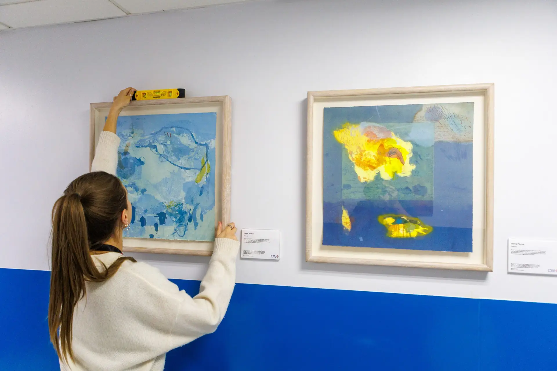 There are two framed artworks on a wall. A woman is installing one of them and checking it is straight with a spirit level
