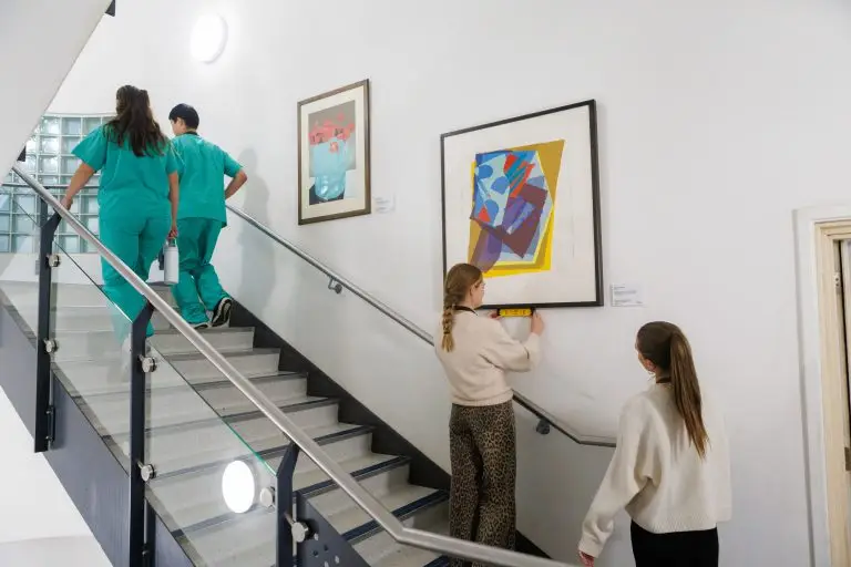 A stairwell with two pieces of art on the wall. There are two members of hospital staff walking up the stairs in scrubs. In front of one of the artworks, one woman is checking it is straight with a spirit level; the other woman looks on.
