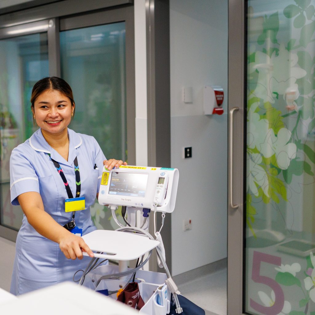 A smiling nurse with hospital equipment in a corridor with private bays behind her. The doors of the bays are decorated with nature-inspired artwork